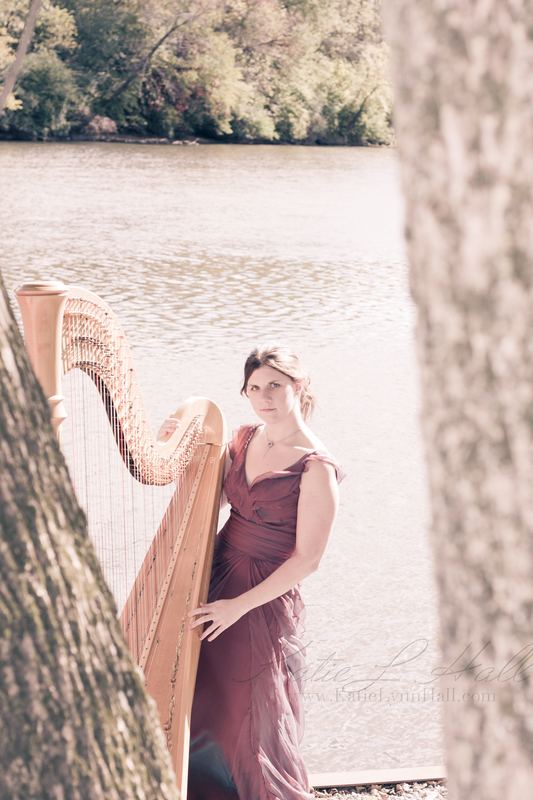 Harpist in a burgundy gown framed by trees beside a river